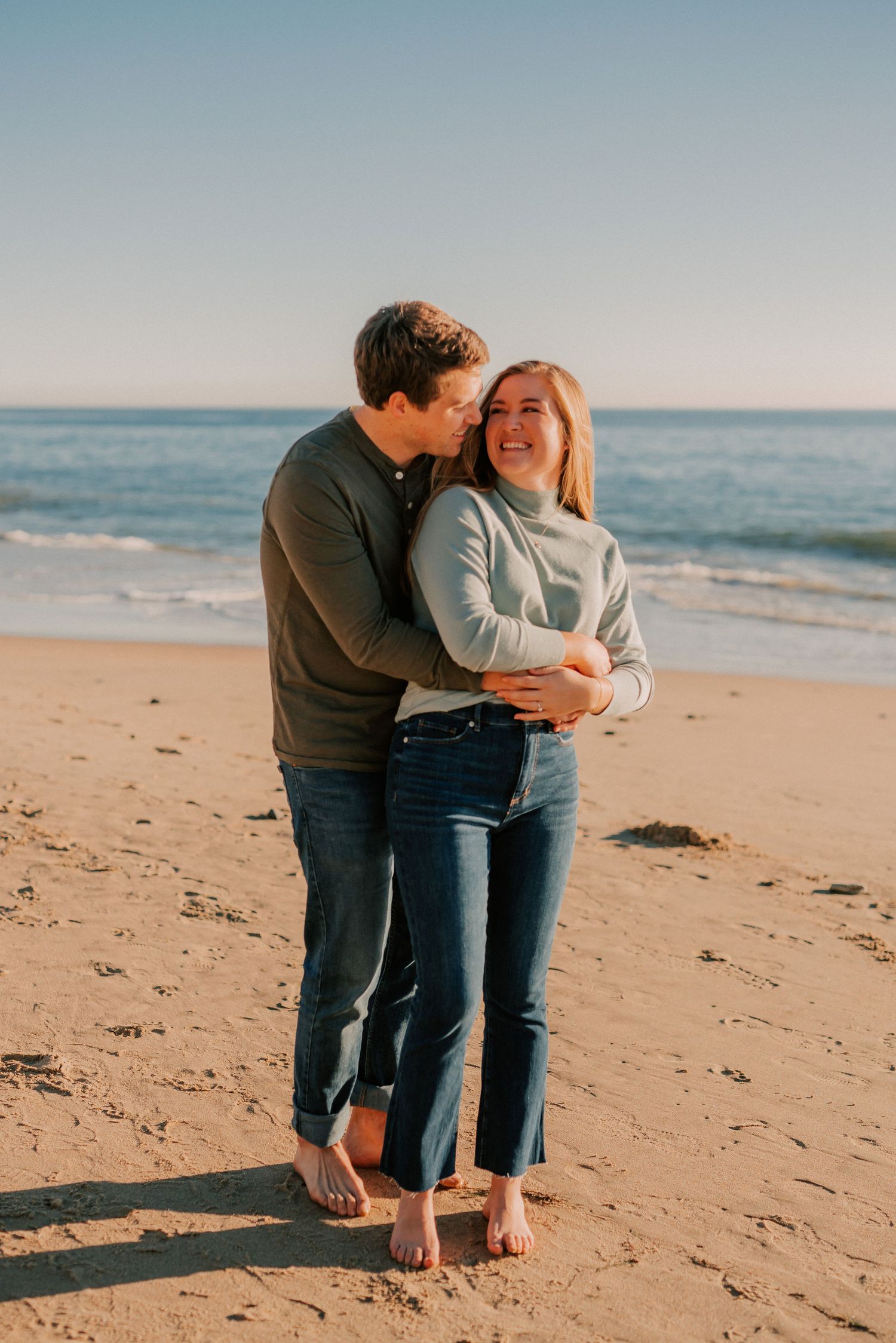 Couple embracing and smiling on a sandy beach at golden hour, wearing casual sweaters and jeans.
