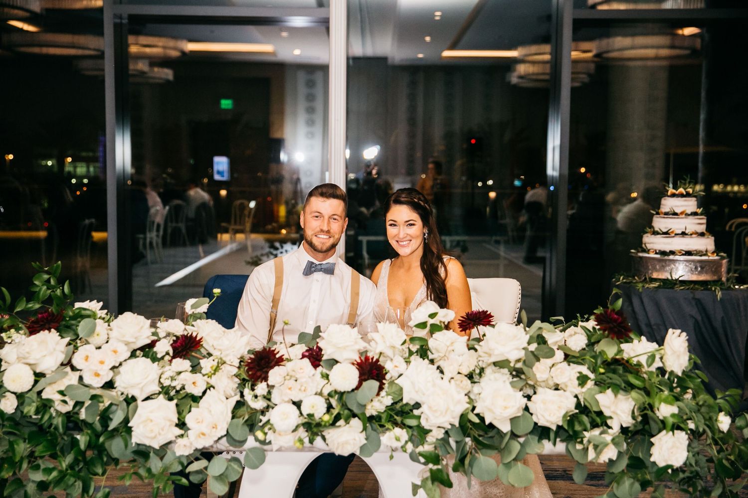 Bride and groom smiling at sweetheart table adorned with white roses and greenery at elegant wedding reception.