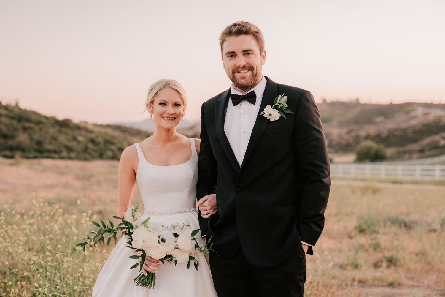 Bride and groom smiling together in a sunlit field, she holds a white bouquet, he wears a black tuxedo.