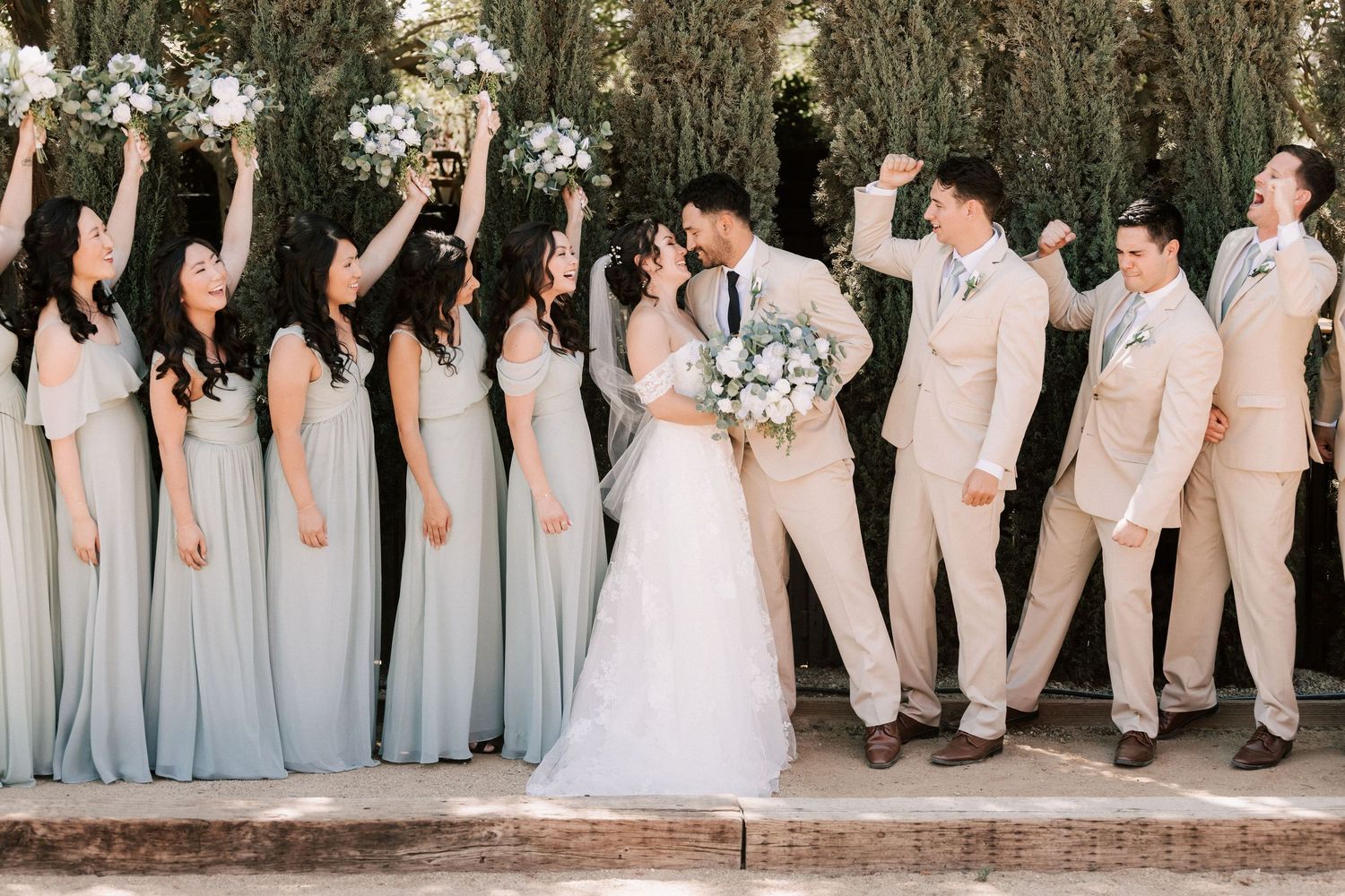 Wedding party celebrating outdoors, bridesmaids in sage green gowns and groomsmen in tan suits cheering as couple kisses.