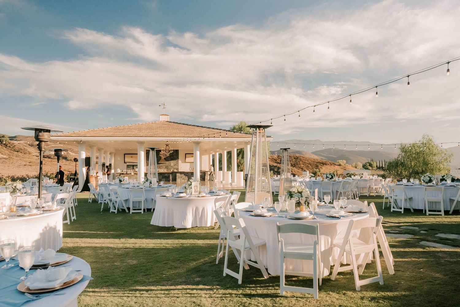 Elegant outdoor wedding reception with white round tables, chairs, string lights, and a gazebo on a lush green lawn.