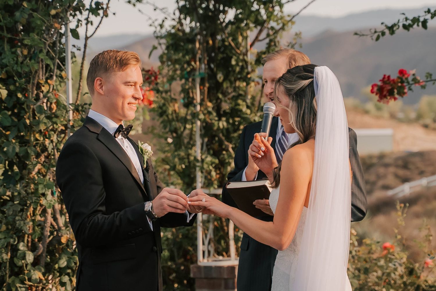 Groom places wedding ring on bride's finger during outdoor ceremony with officiant and floral arch backdrop.