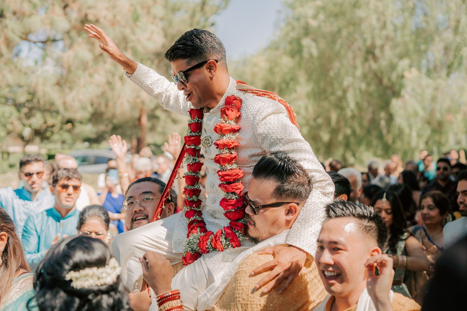 Groom lifted on shoulders by groomsmen while waving to crowd during Indian wedding baraat celebration.