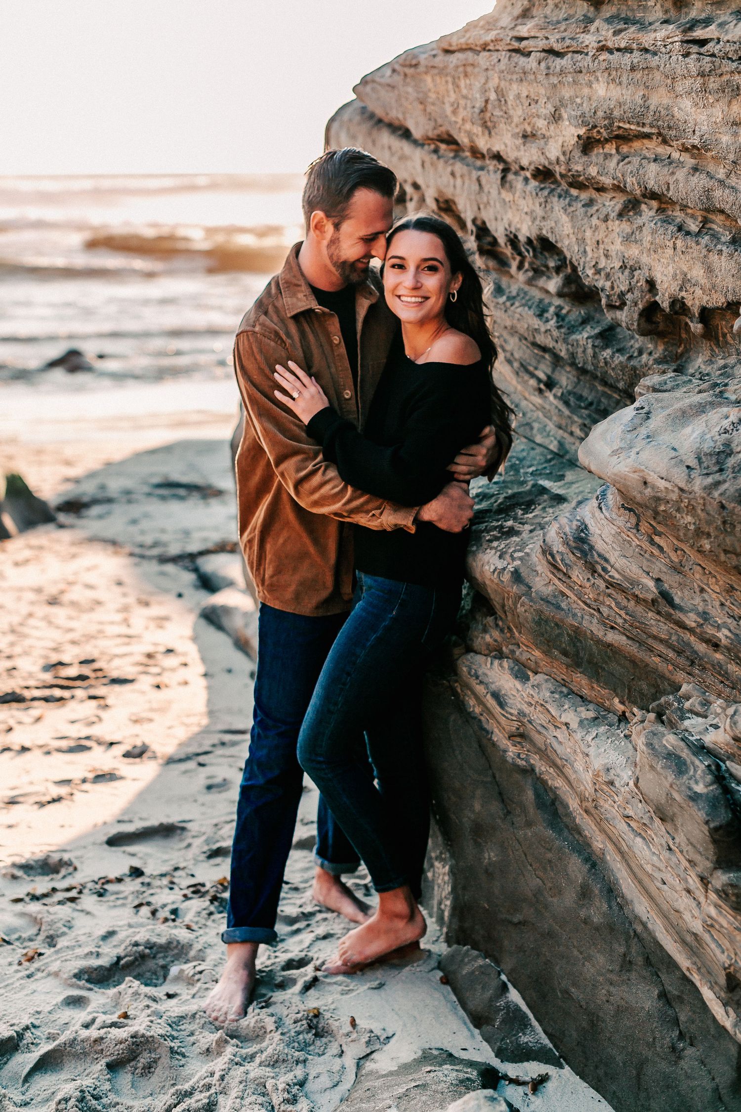 Couple embracing against layered sandstone cliffs on a beach, smiling warmly at golden hour.
