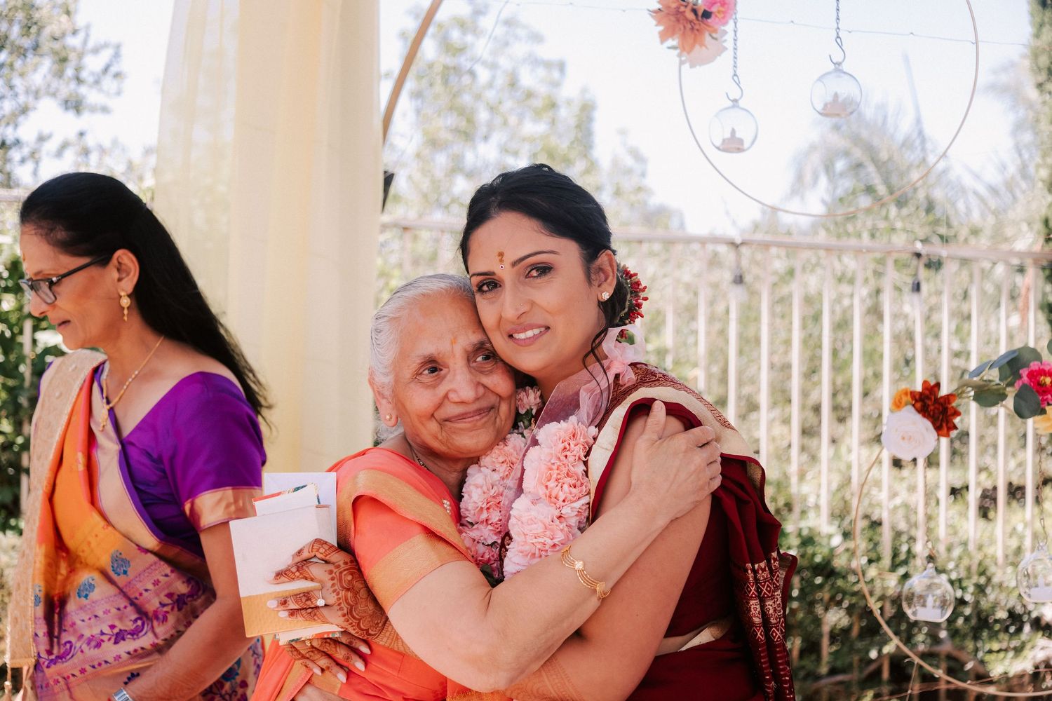 Bride in red saree sharing a warm embrace with elderly woman at an Indian wedding celebration.