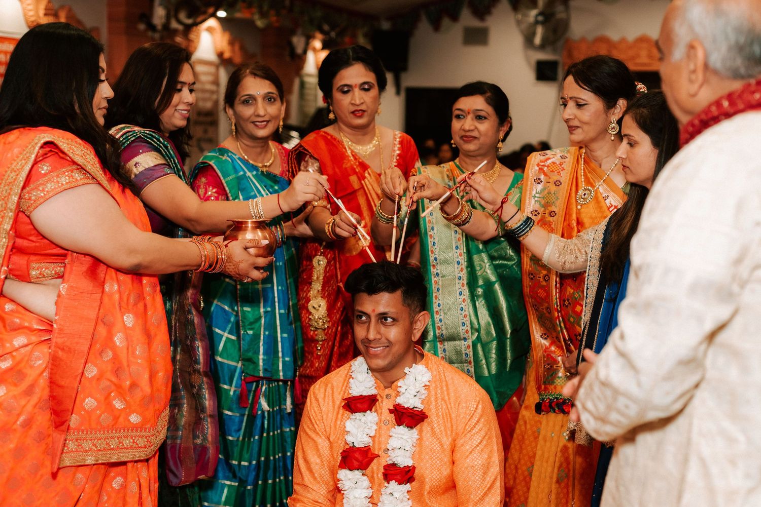 Groom in orange kurta with floral garland surrounded by women in colorful sarees during Hindu wedding rituals.