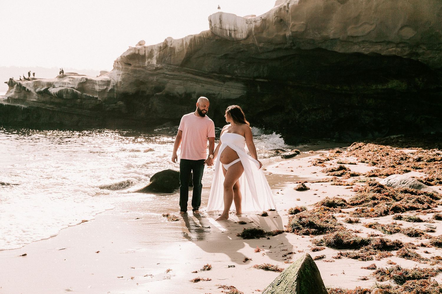 Couple walking on sunlit beach, pregnant woman in white flowing dress, man in pink shirt.