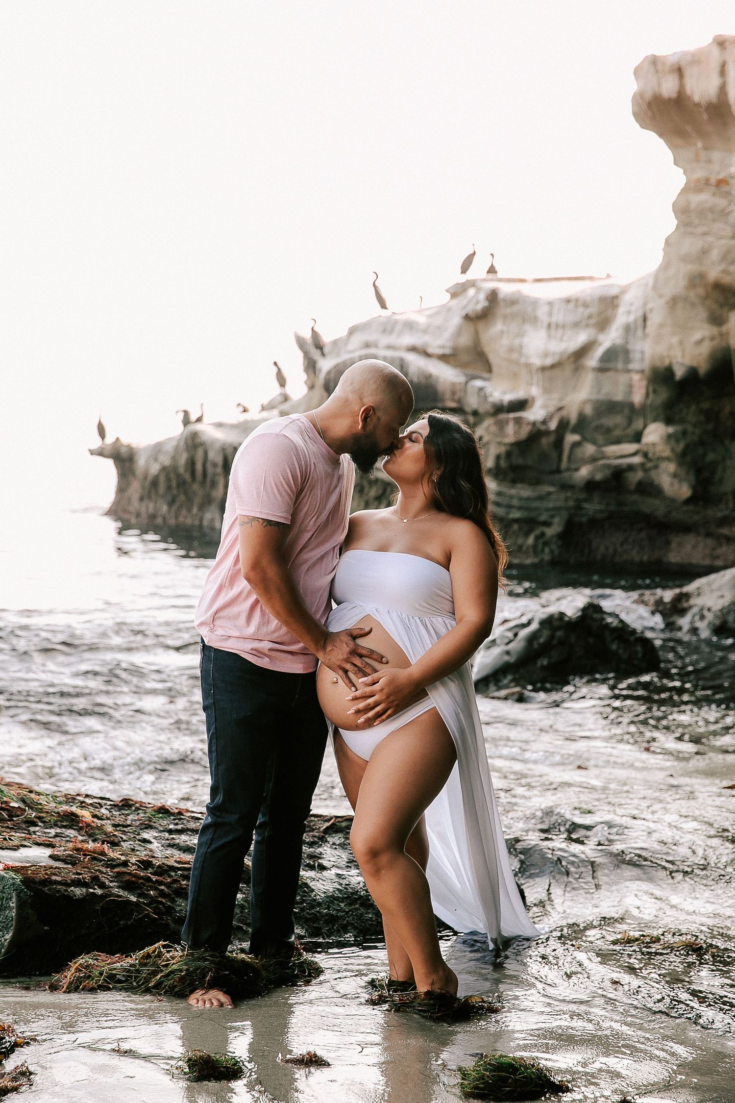 Couple sharing a tender kiss near rocky shoreline, pregnant woman in white flowing maternity gown.