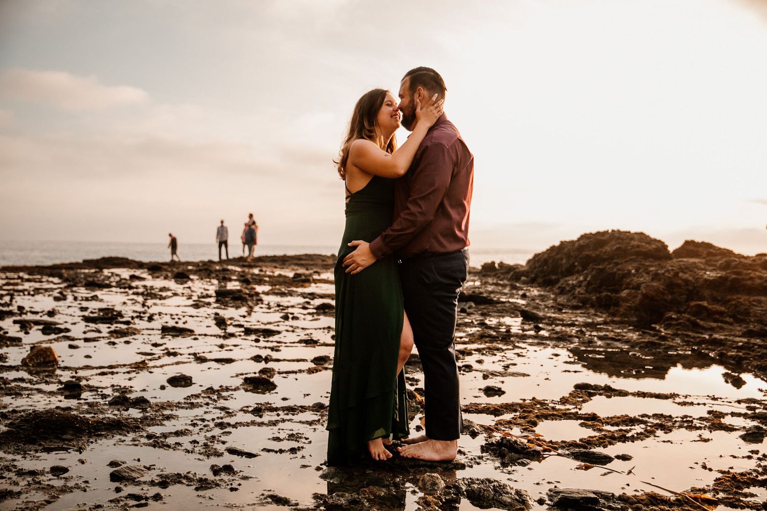 Couple sharing a romantic moment at sunset on a rocky beach, with silhouetted figures in the background.