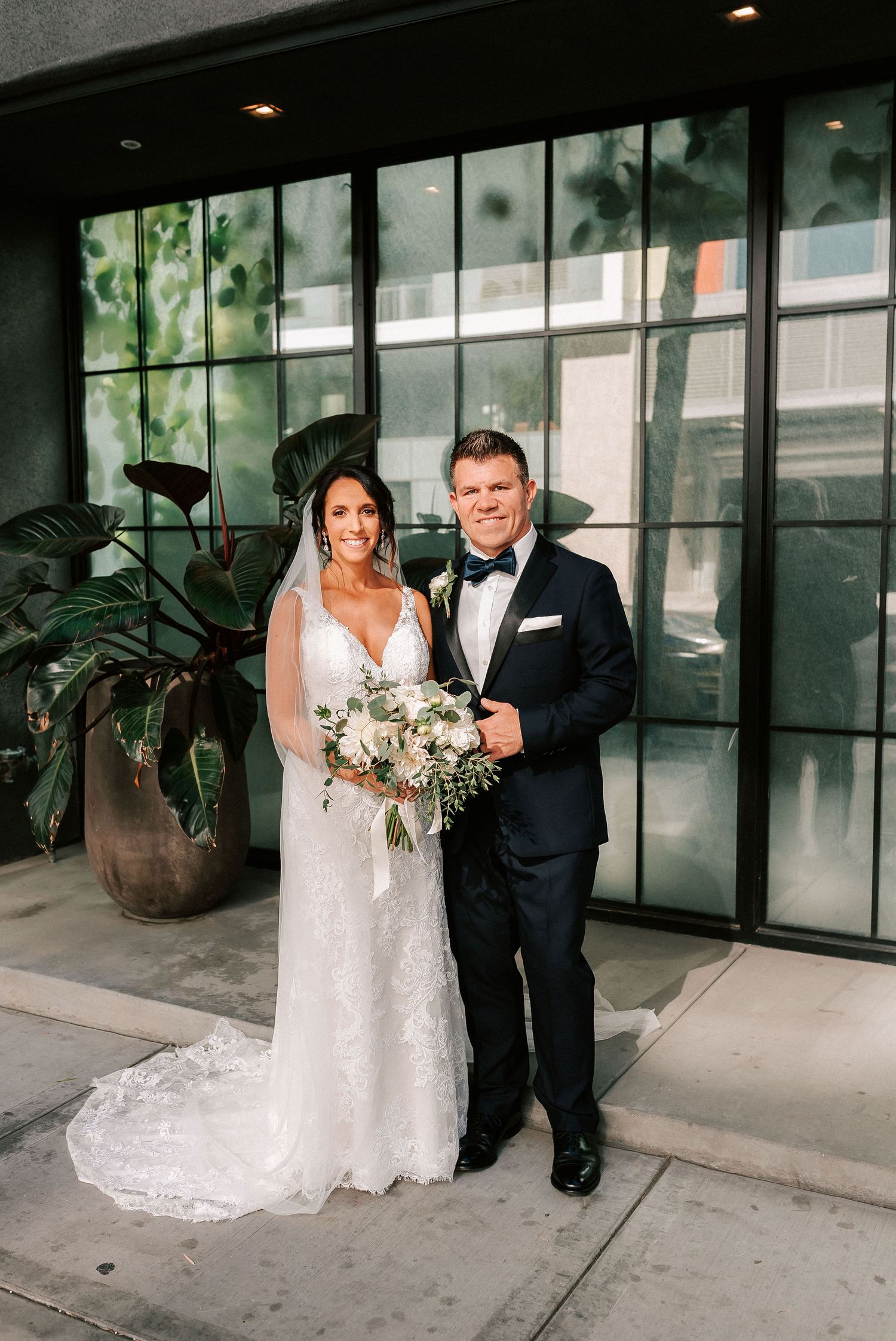 Bride in lace gown and groom in navy tuxedo pose together holding white floral bouquet outside modern venue.