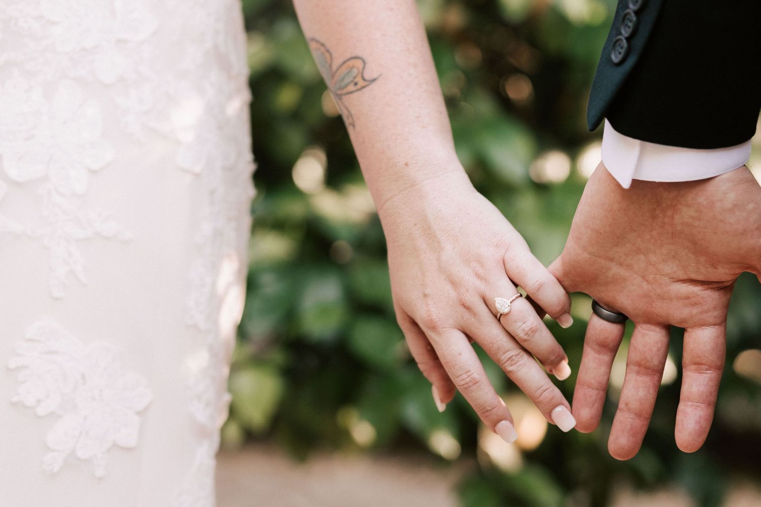 Bride and groom holding hands showing wedding rings, with lace wedding dress and green foliage background.