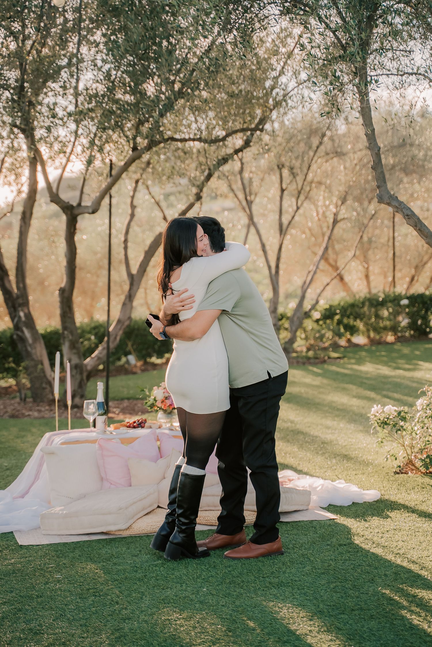 Couple embracing in a romantic outdoor garden picnic setup at golden hour with lush greenery and trees.