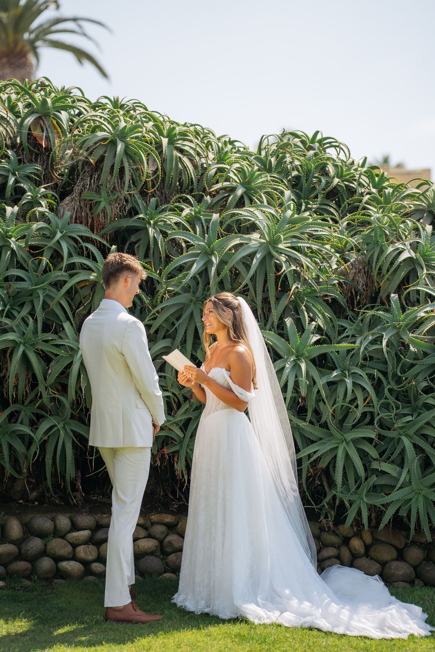 Bride reads vows to groom outdoors in lush tropical garden setting, wearing elegant white off-shoulder gown with veil.