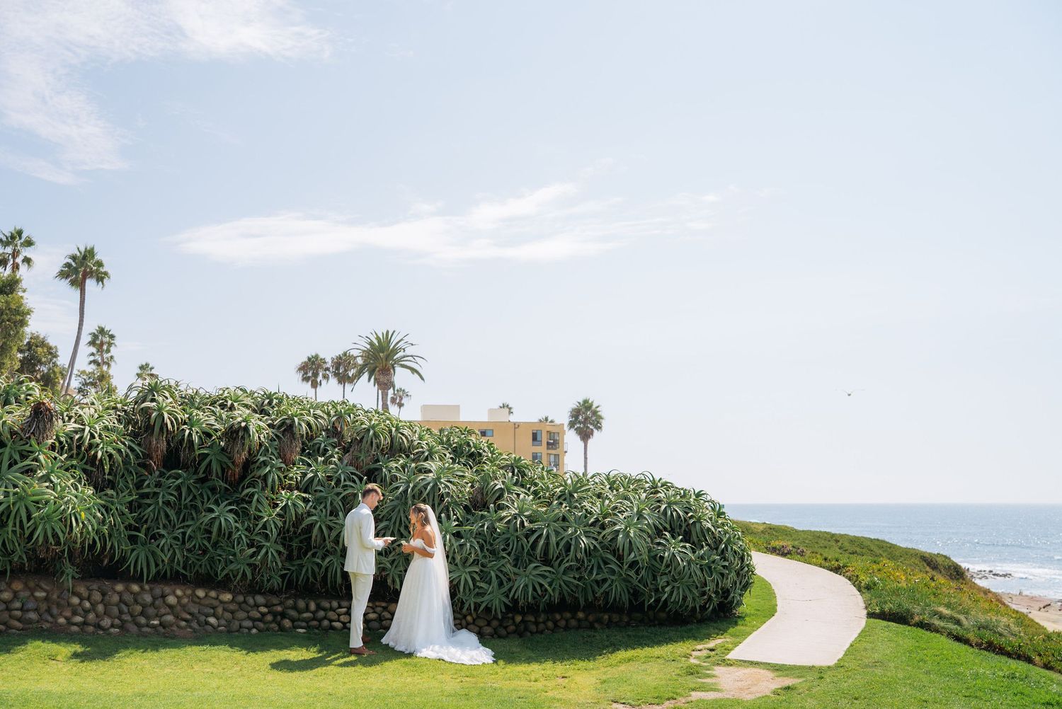 Bride and groom stand together on lush green lawn with palm trees and ocean view behind them.