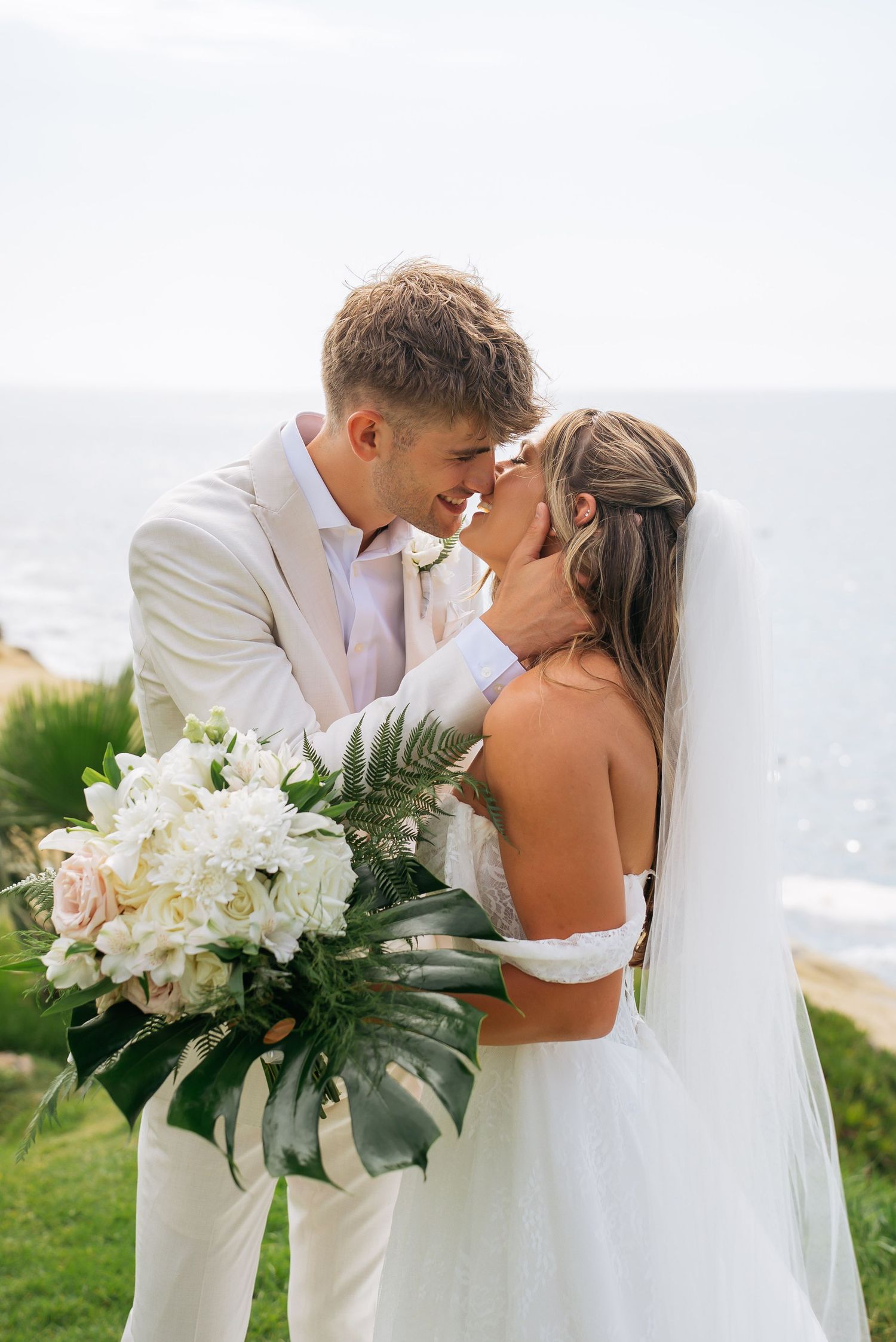 Bride and groom share a romantic forehead touch holding white floral bouquet with ocean backdrop.