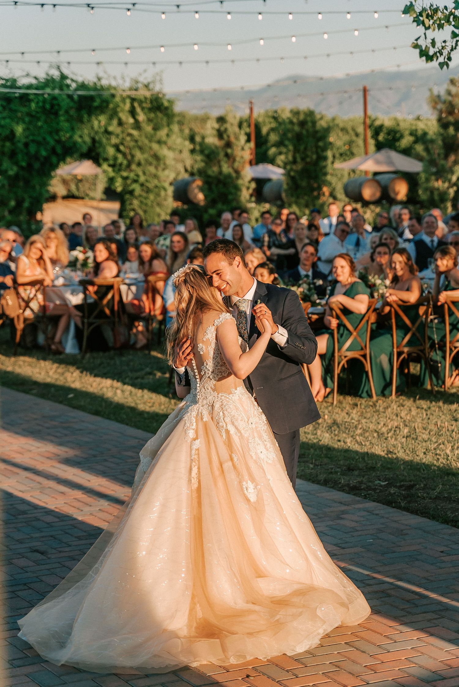 Bride and groom share first dance at outdoor wedding reception with string lights and mountain views at golden hour.