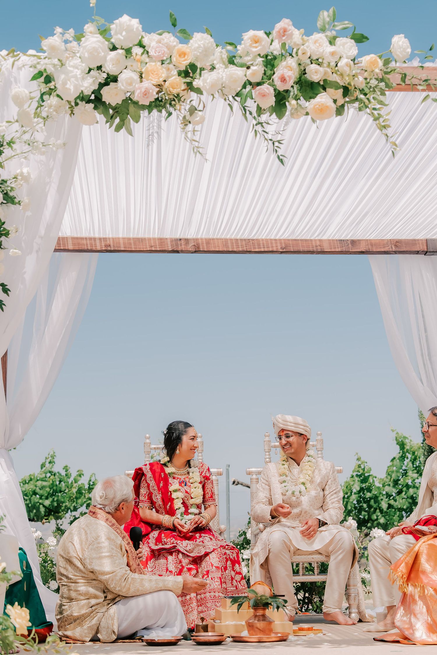 Indian bride in red lehenga and groom seated during Hindu wedding ceremony under floral mandap.
