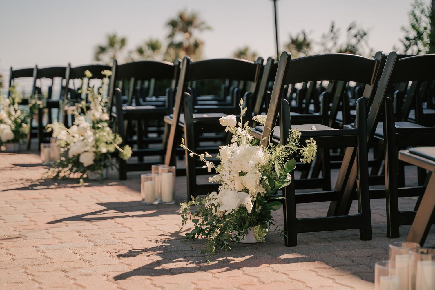 Rows of black folding chairs line an outdoor wedding ceremony aisle with white floral arrangements, candles, and palm trees in the backgroun