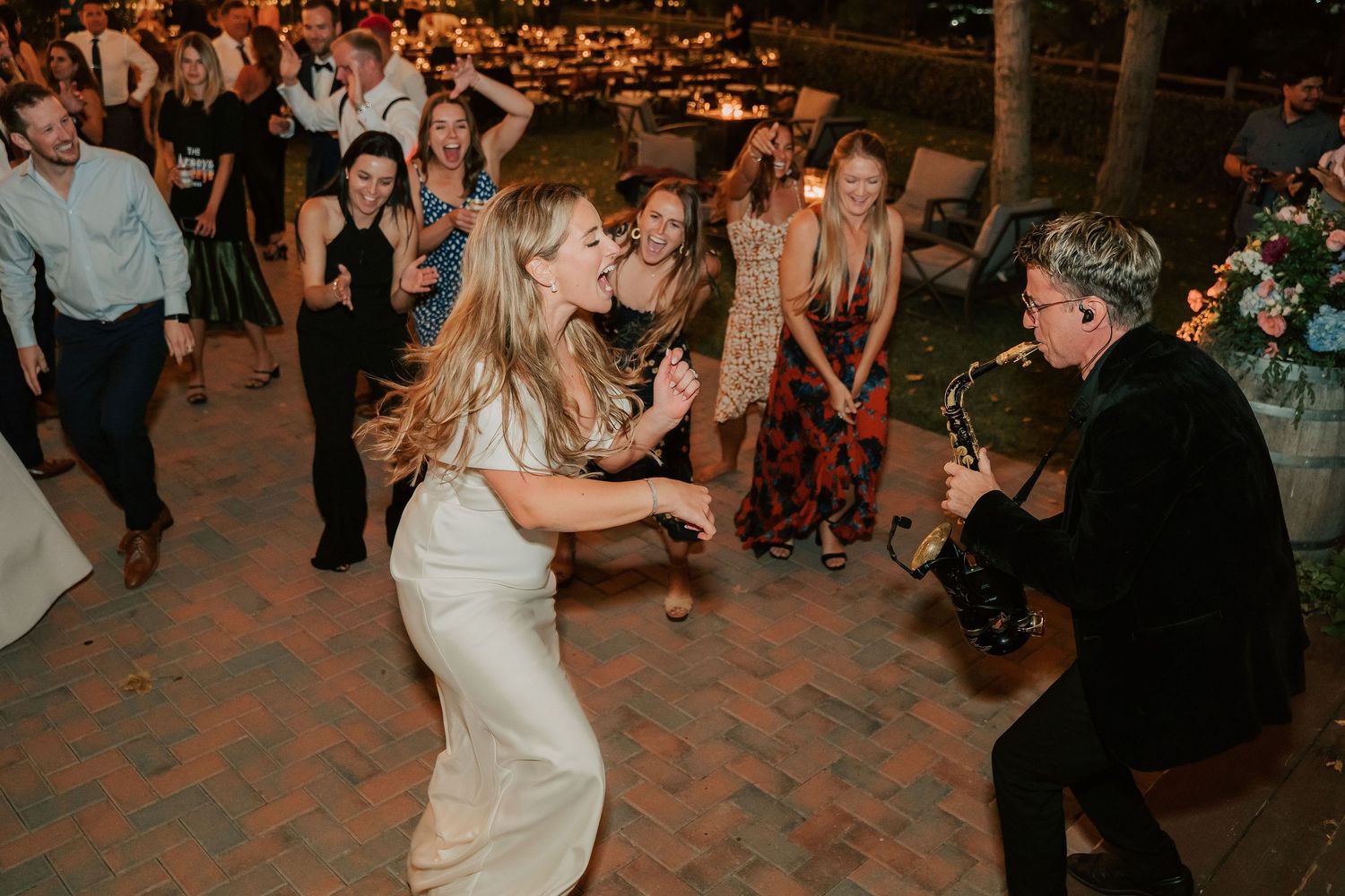 Bride in white dress dancing with guitarist at outdoor wedding reception while guests cheer in background.