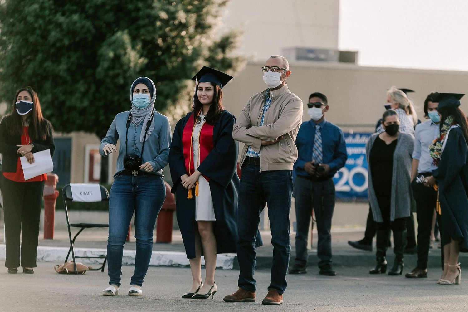Graduate in cap and gown stands with family at outdoor graduation ceremony, all wearing face masks.