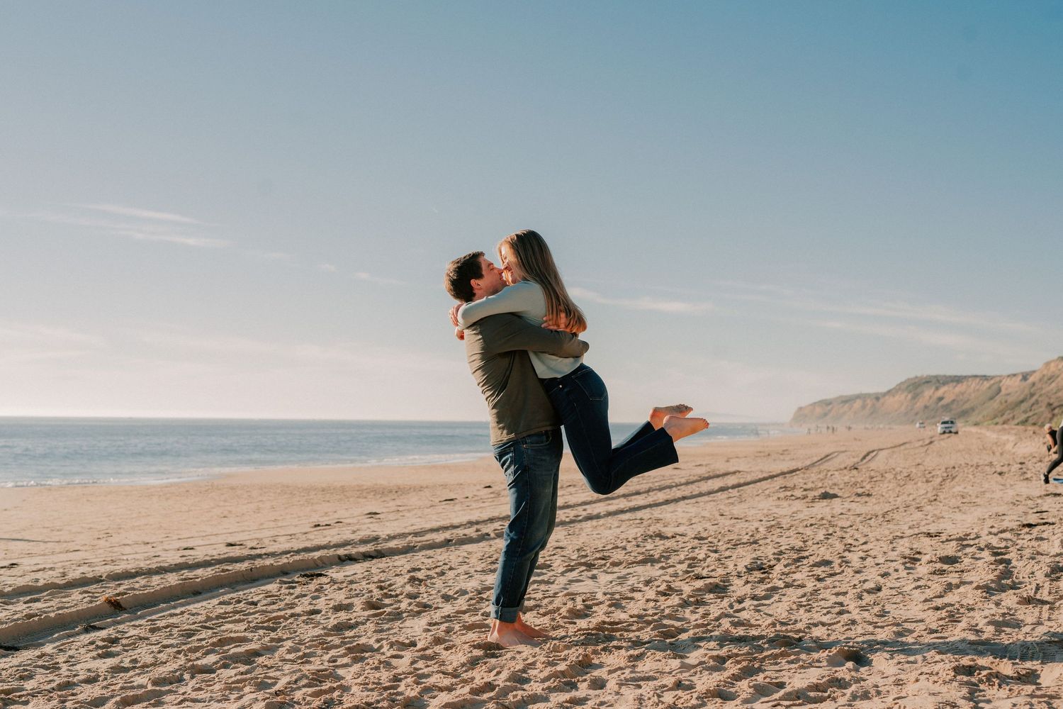 Couple embracing and kissing on a sunny sandy beach with ocean waves and cliffs in the background.