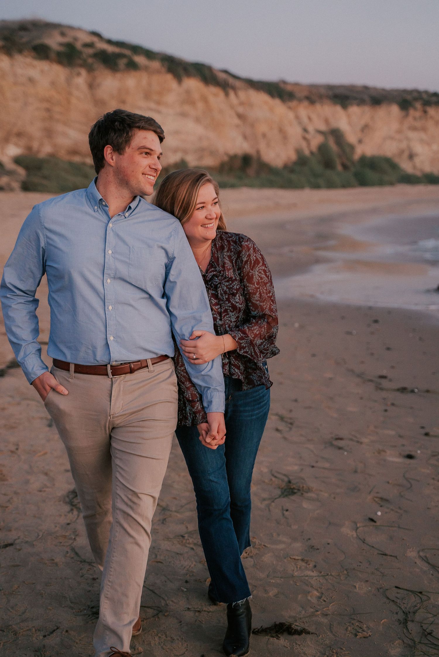 Couple walking hand-in-hand on a sandy beach at sunset with cliffs in the background.