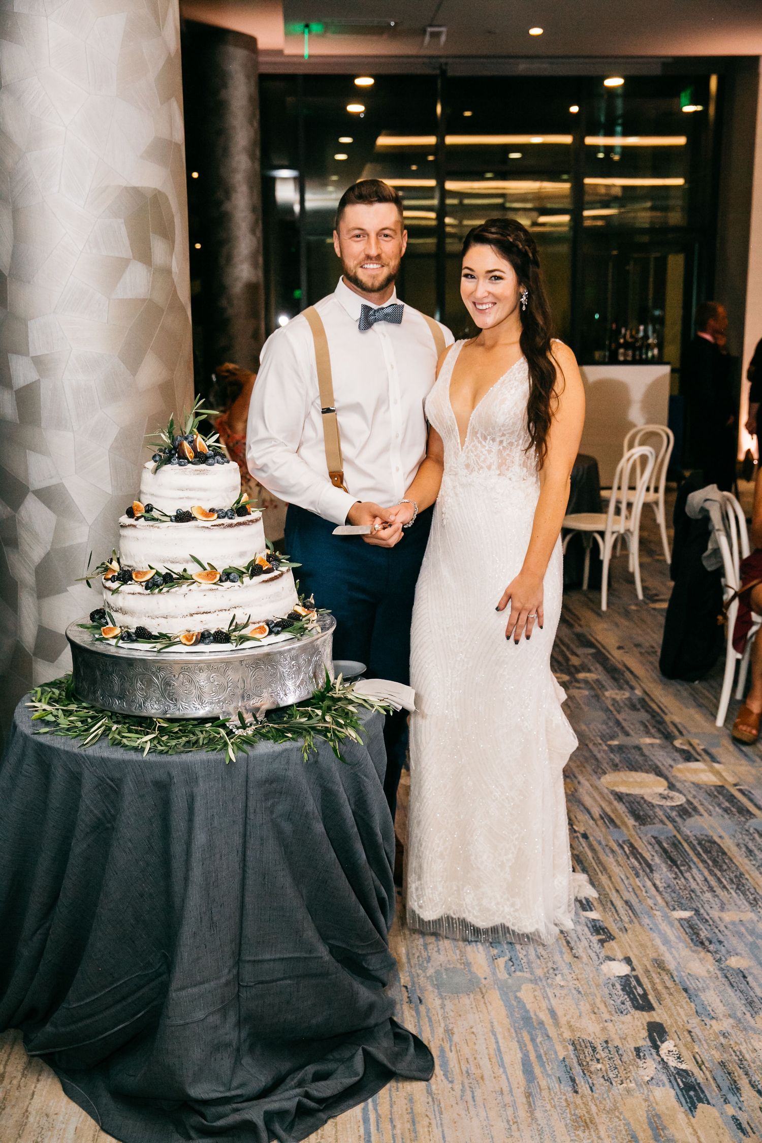 Bride and groom smiling together while cutting their four-tier wedding cake decorated with greenery and flowers.