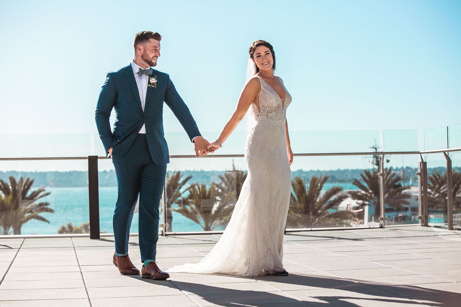 Bride and groom holding hands on a sunny terrace with palm trees and ocean views on their wedding day.