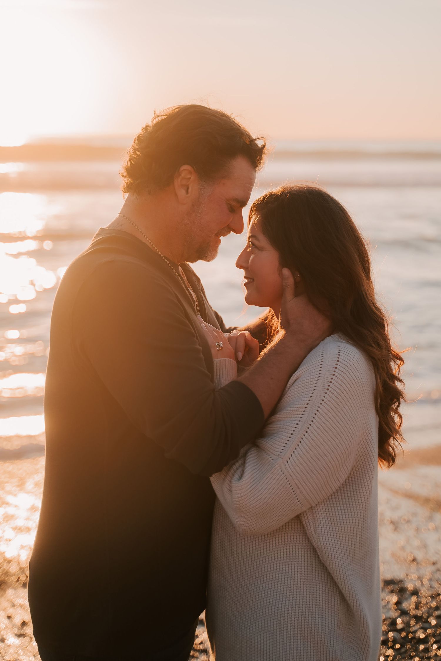 Couple embracing face to face on a beach at golden hour sunset, smiling as warm light glows over the ocean behind them.
