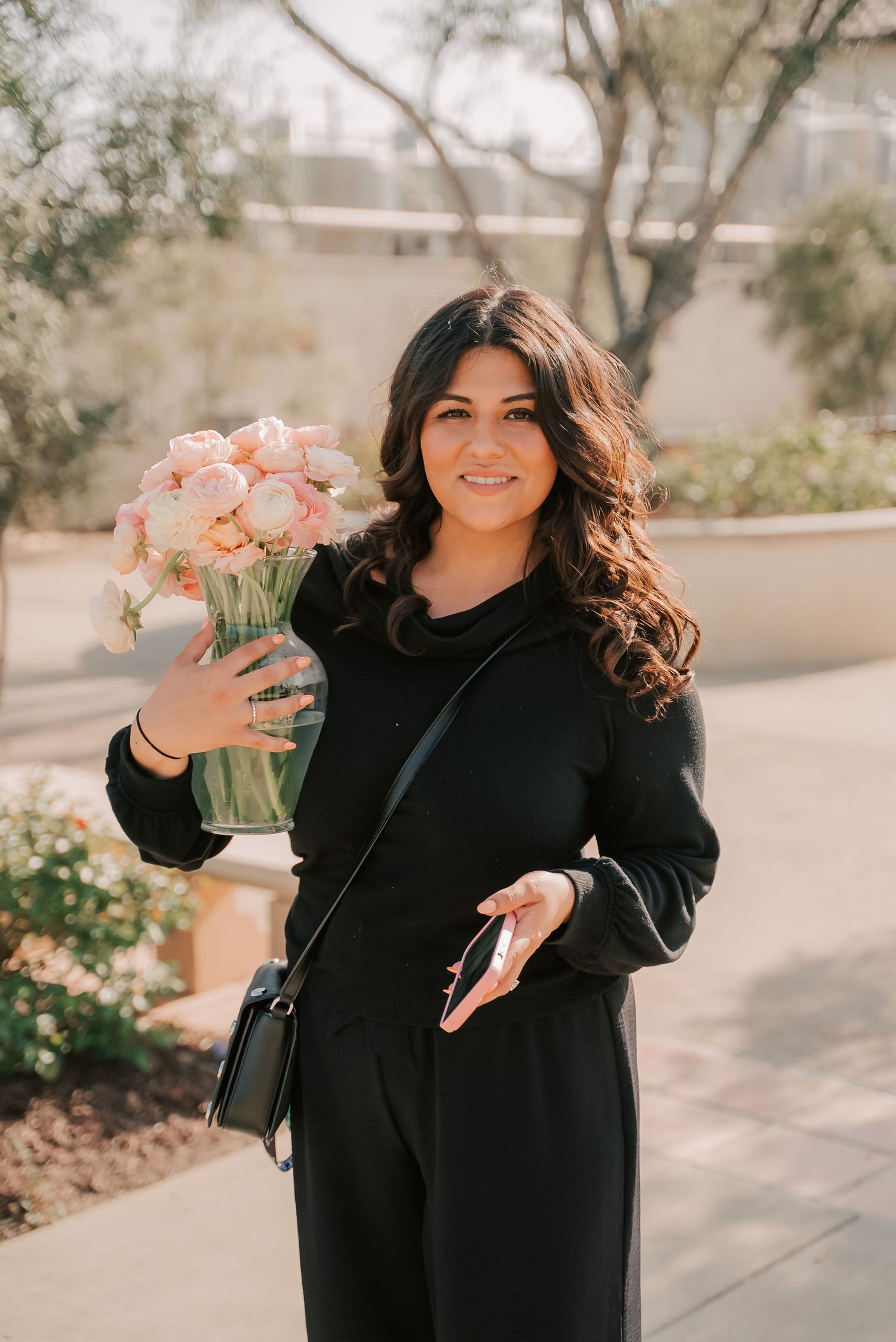 Woman in black outfit holding pink floral arrangement in glass vase, smiling outdoors in garden setting.