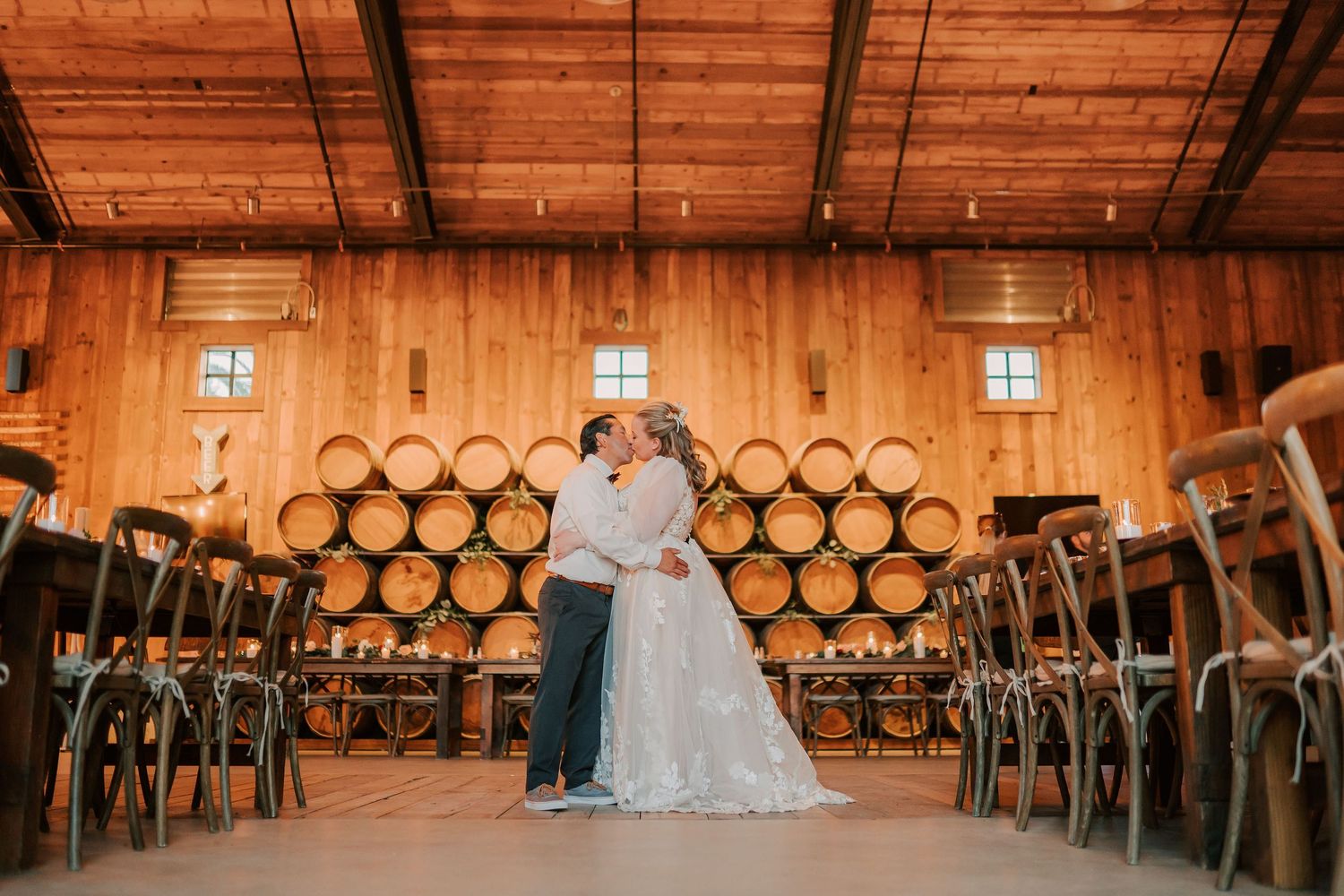 Bride and groom embrace in rustic winery venue with wooden barrel wall backdrop and elegant cross-back chairs.