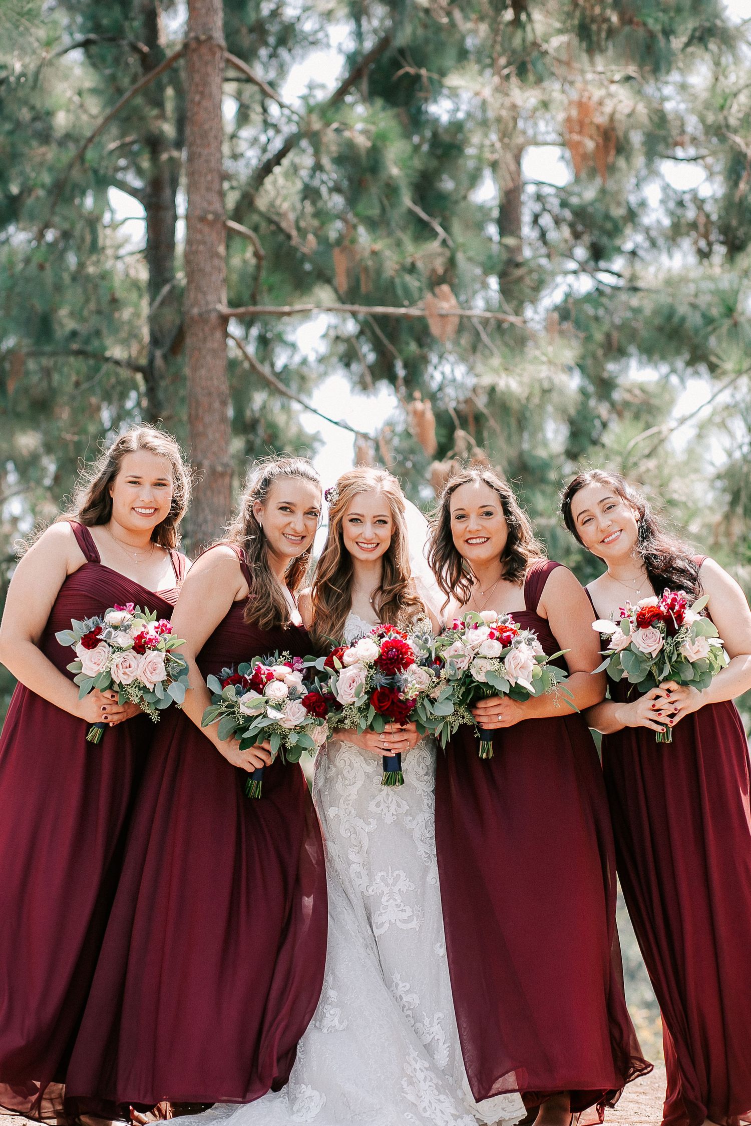 Bride in white lace gown poses with four bridesmaids in burgundy dresses holding floral bouquets outdoors.