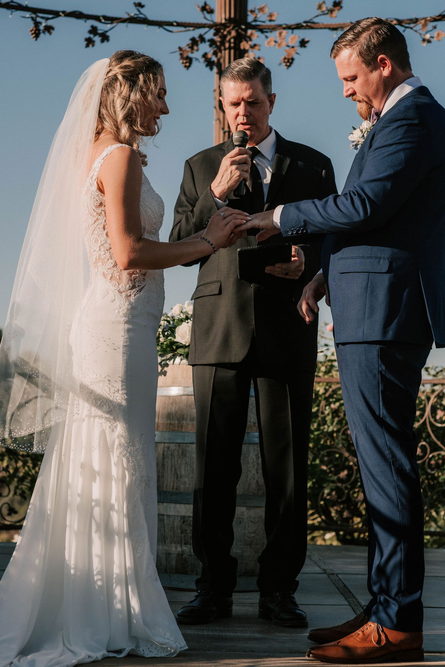 Bride and groom exchanging rings during outdoor wedding ceremony with officiant speaking into microphone.