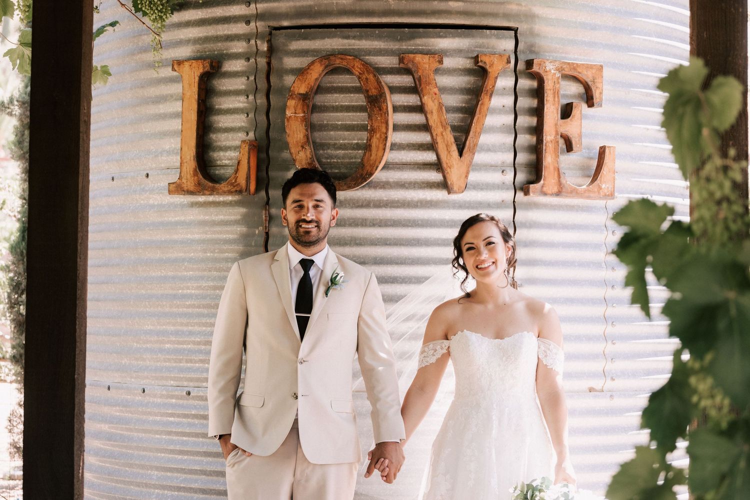 A bride and groom hold hands smiling before a rustic corrugated metal wall with large LOVE letters at their wedding.