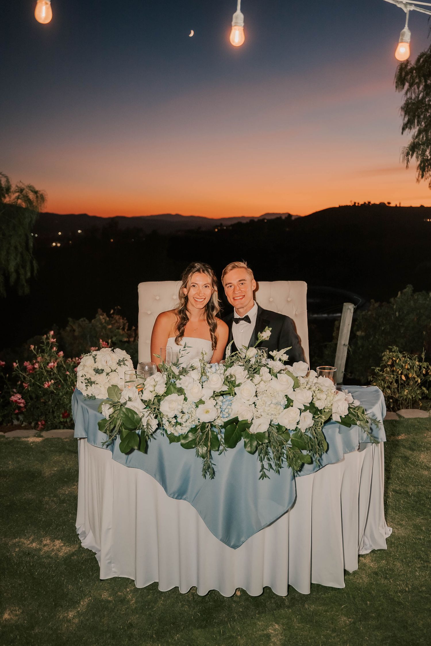 Bride and groom seated at sweetheart table with white floral arrangements at outdoor wedding reception during sunset.