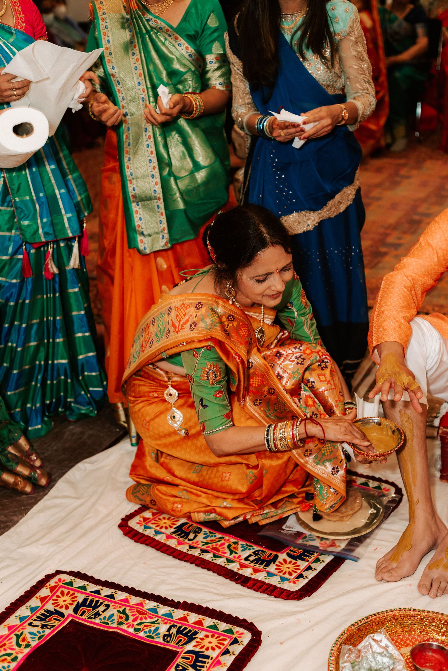 A woman in an orange and green saree performing a traditional Hindu wedding ritual while seated on the floor.