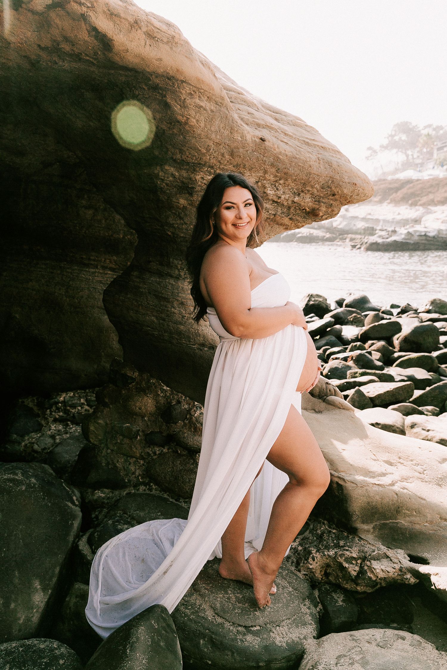Smiling pregnant woman in white strapless maternity gown leaning against large rock at beach sunset.