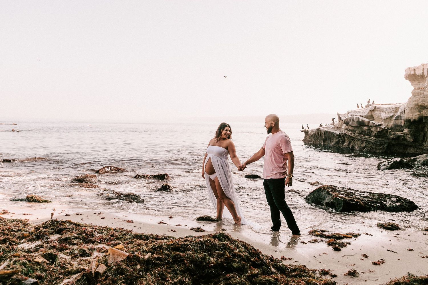 Expecting couple holding hands walking along a rocky beach at sunset during outdoor maternity session.
