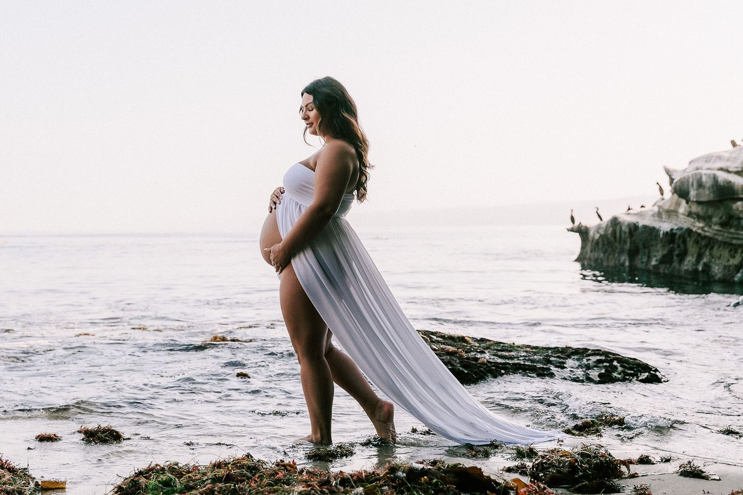 Pregnant woman in sheer white maternity gown standing on rocky beach at sunset, cradling baby bump.