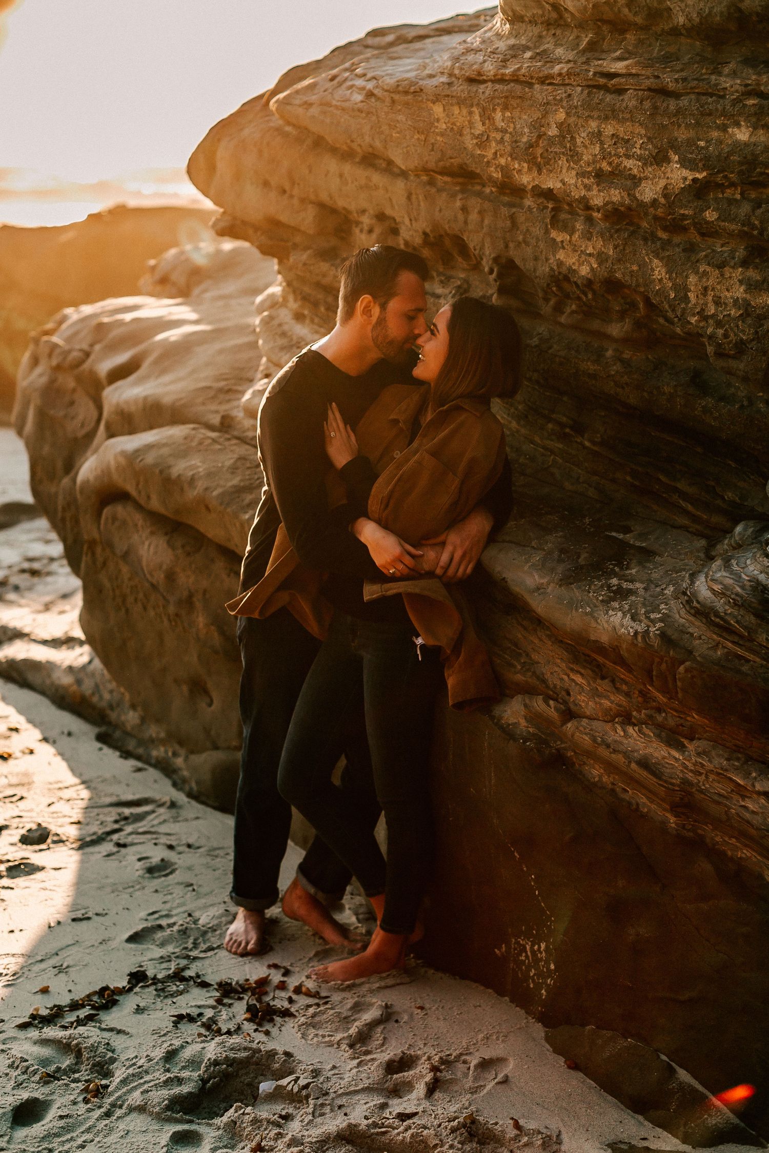 Couple sharing an intimate moment leaning against golden sandstone cliffs on a beach at sunset.