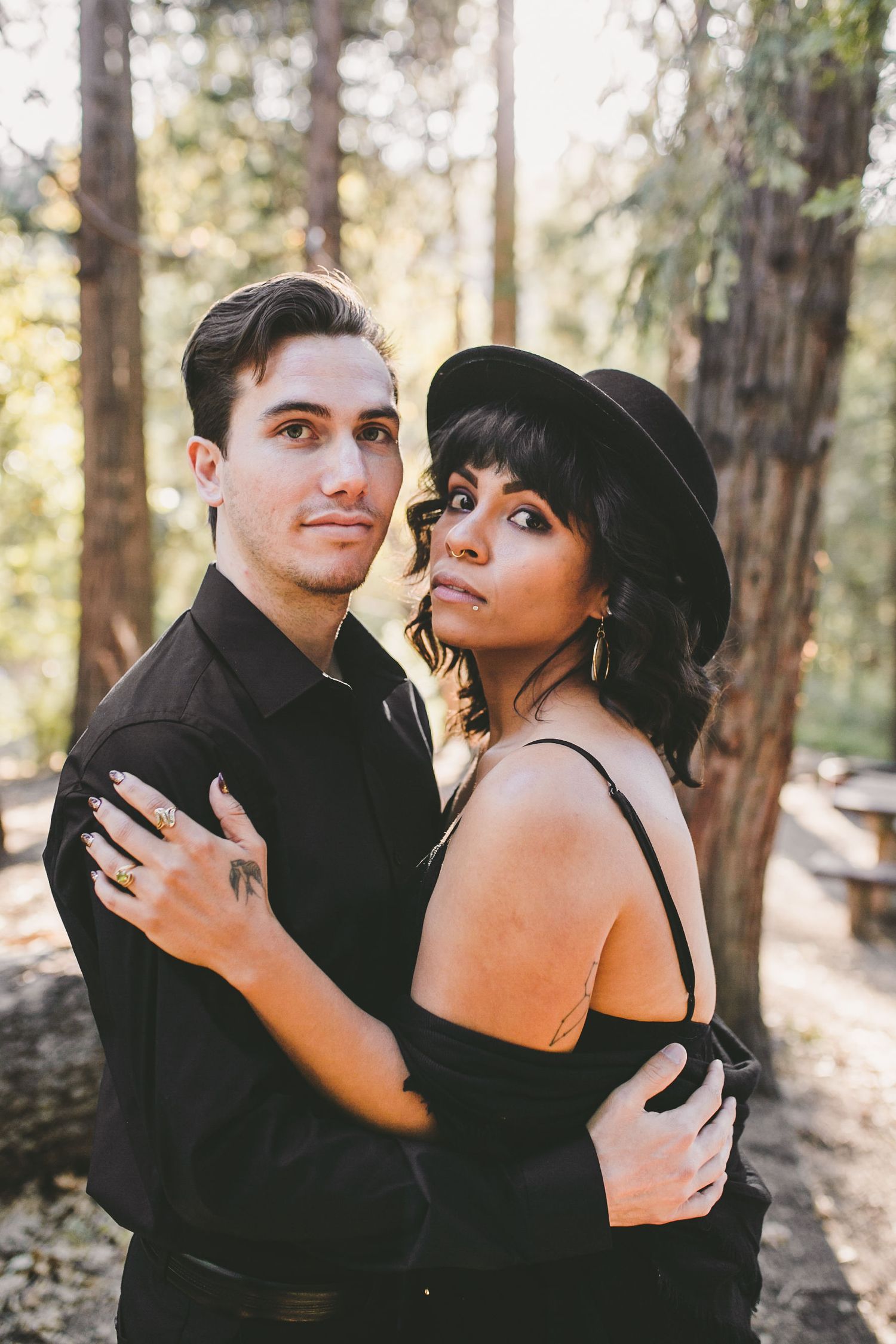 A couple in matching black outfits embraces in a sunlit forest, the woman wearing a black hat and gold earrings.