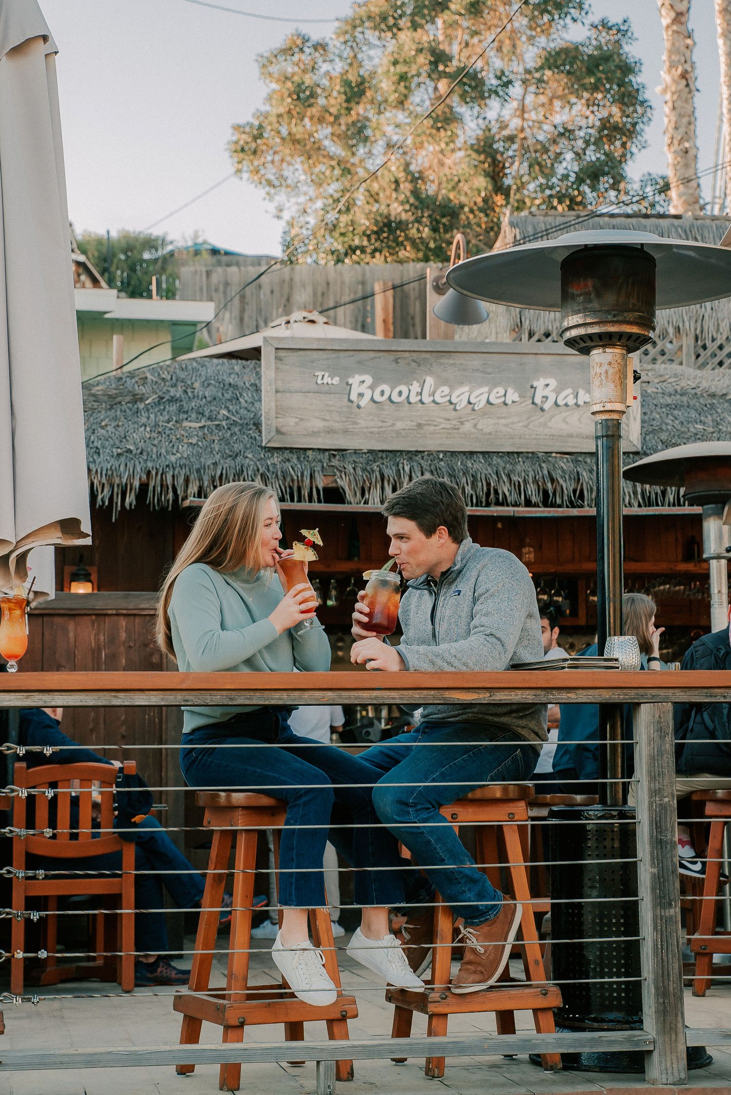 Couple enjoying cocktails at outdoor patio of The Bootlegger Bar, sitting on wooden stools in matching blue sweaters.