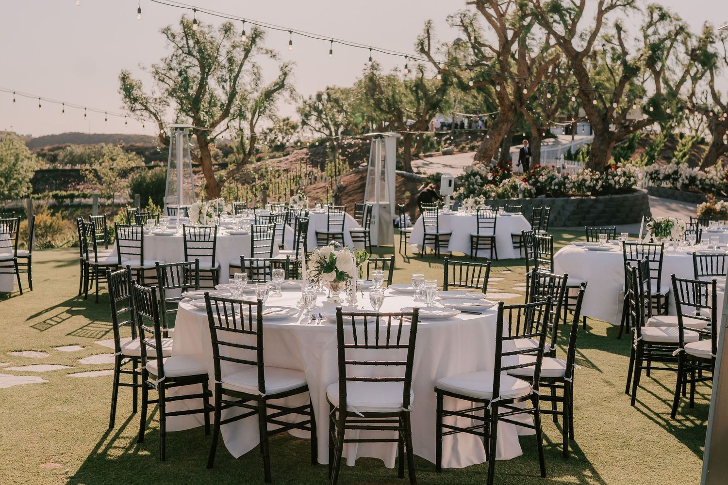 Elegant outdoor wedding reception with round tables, black Chiavari chairs, and string lights among trees.