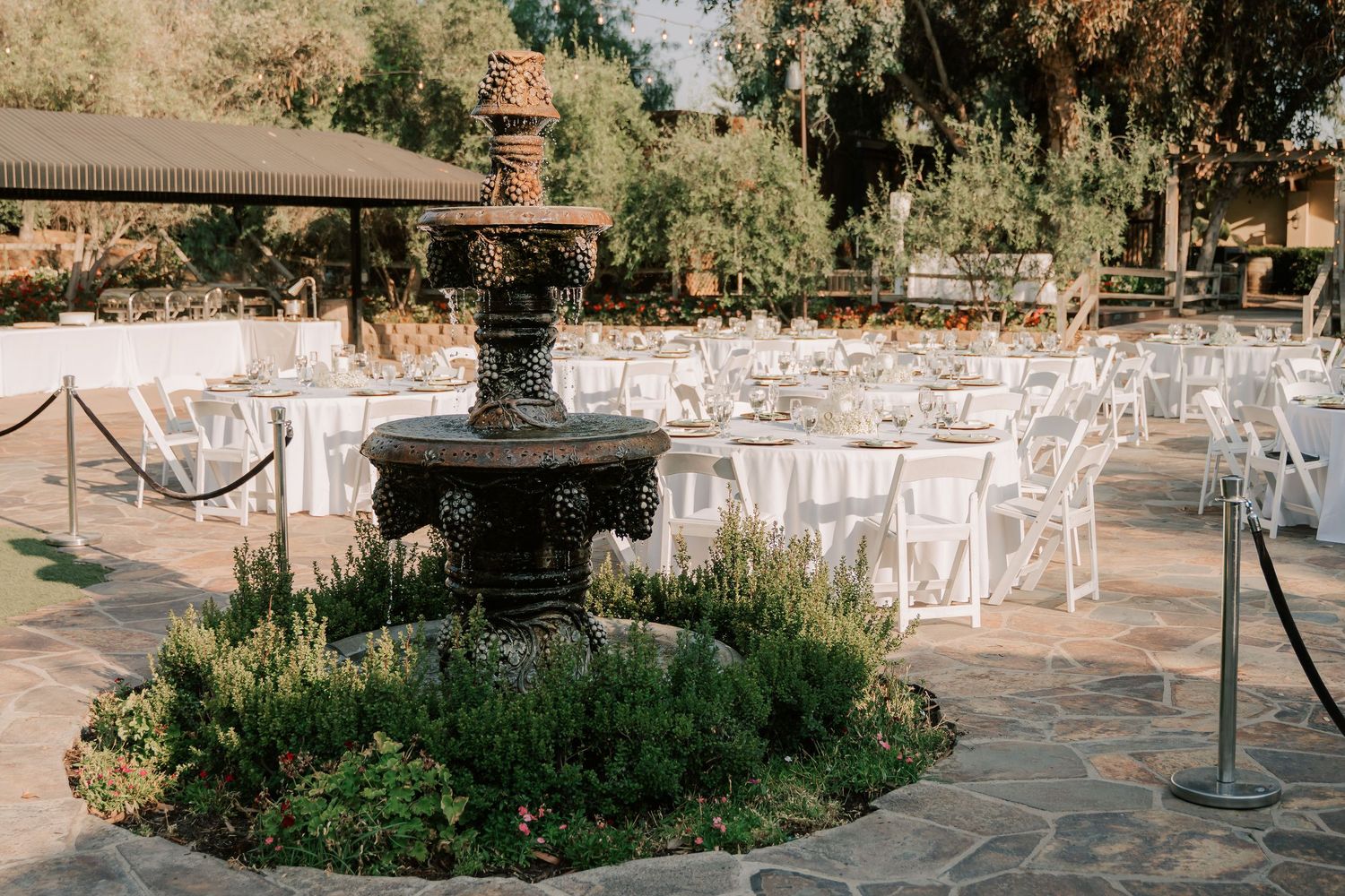 Elegant outdoor wedding reception with white chairs, round tables, and a decorative stone fountain centerpiece surrounded by lush greenery.