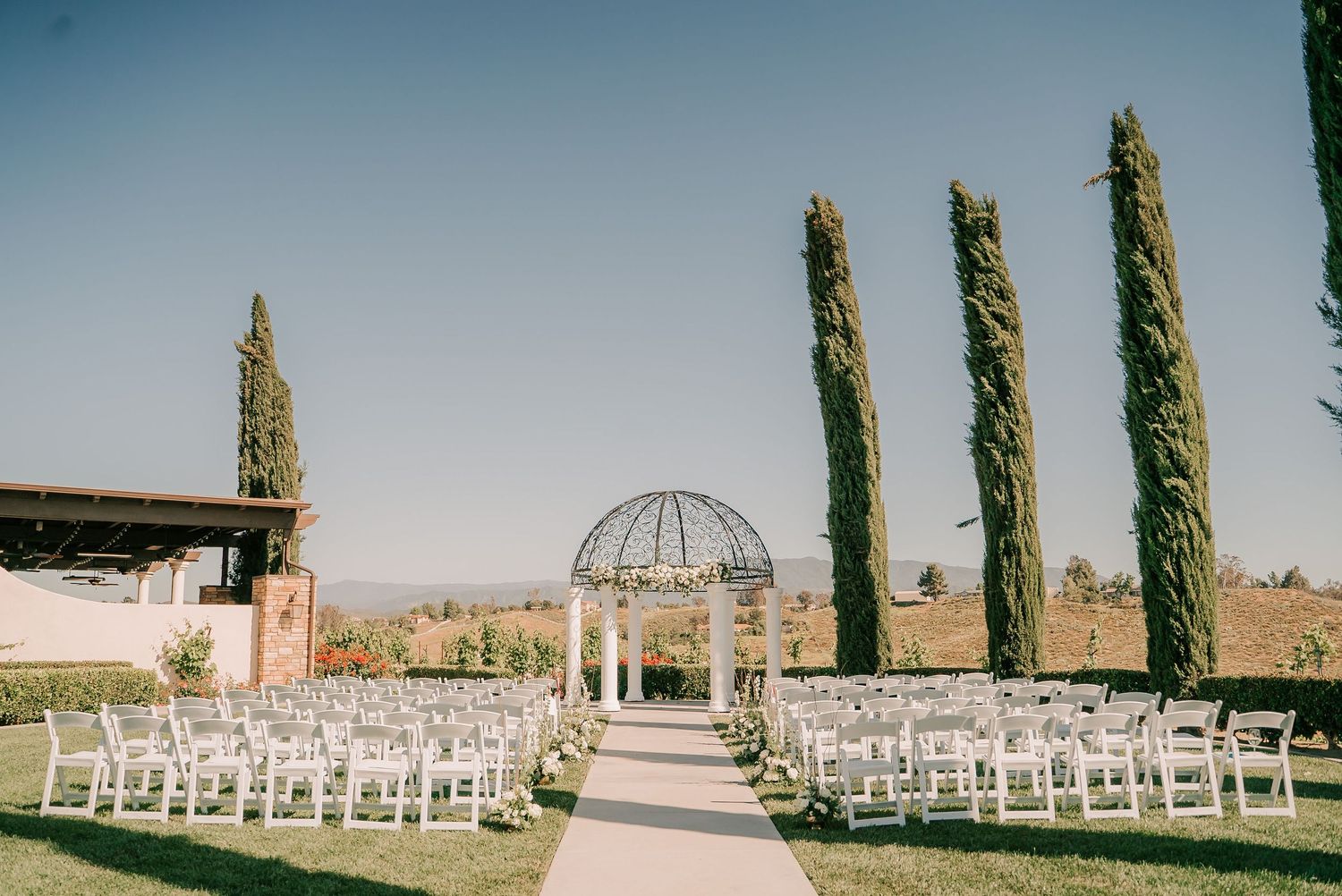 Outdoor wedding ceremony setup with white chairs, elegant gazebo, and tall cypress trees under a clear blue sky.