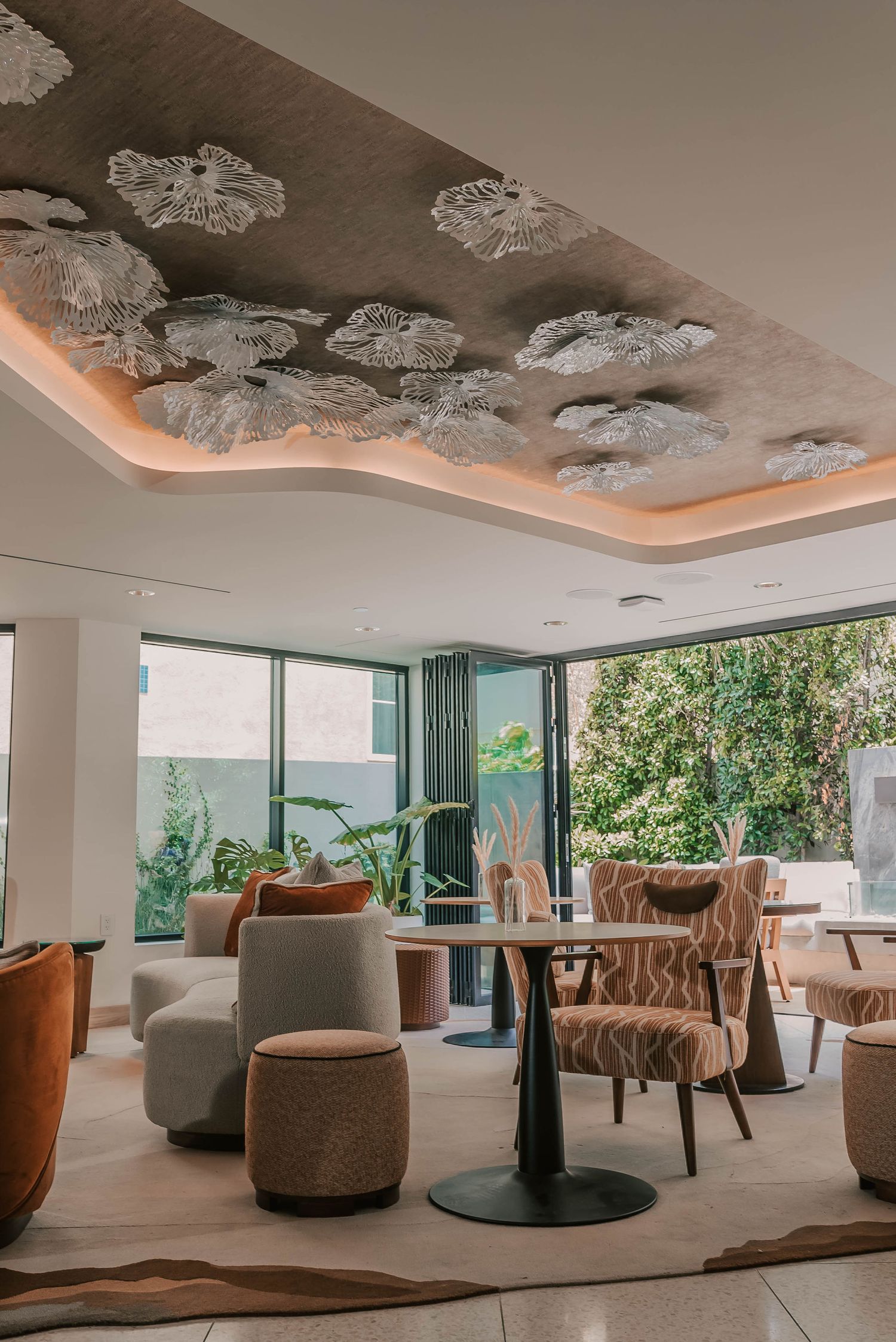 Elegant hotel lounge with floral ceiling art, warm lighting, stylish chairs, and floor-to-ceiling windows overlooking lush greenery.