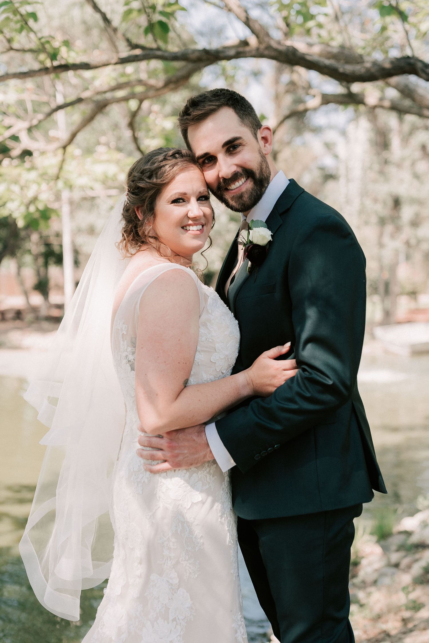 Bride in lace gown and veil smiles with groom in dark suit outdoors near trees and water.