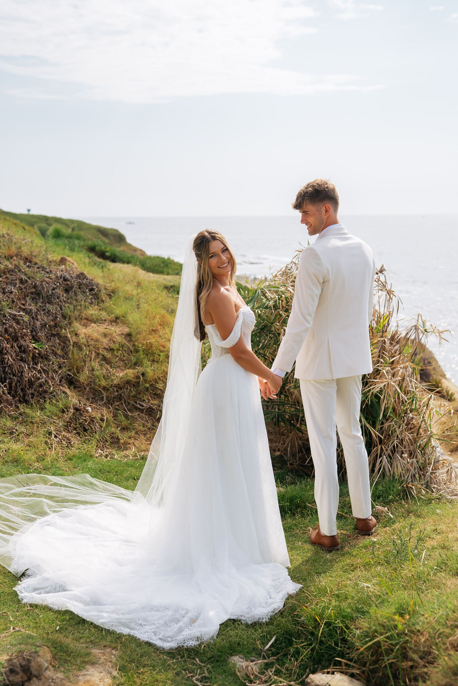 Bride in flowing white gown and veil holds groom's hand while smiling near oceanside greenery.