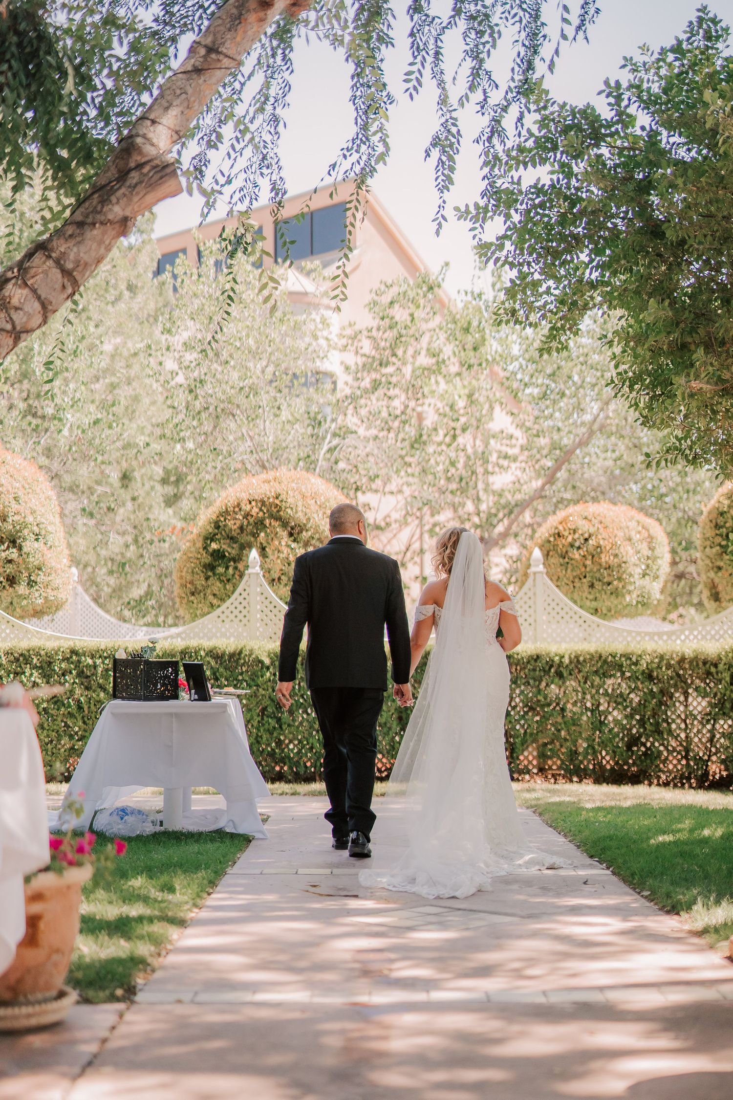 Bride and groom walking hand in hand down an outdoor garden pathway surrounded by lush greenery at a wedding ceremony.