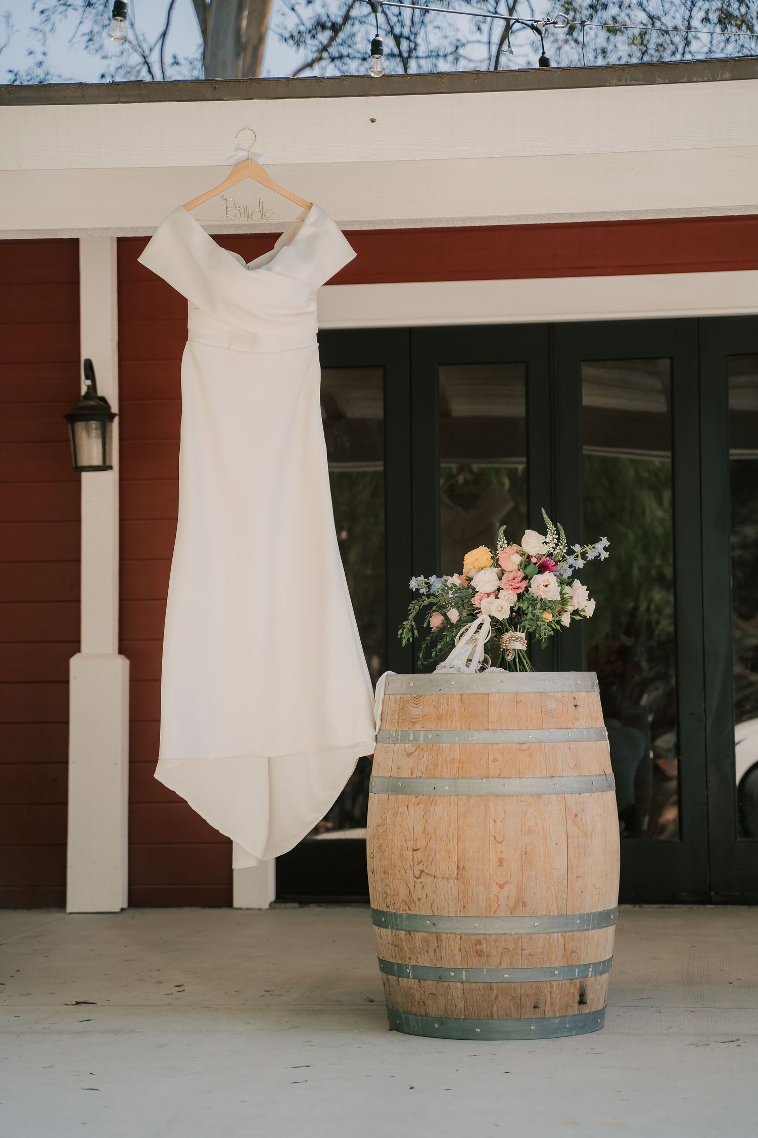 White wedding dress on hanger above floral bouquet on rustic wood barrel outside red barn venue.